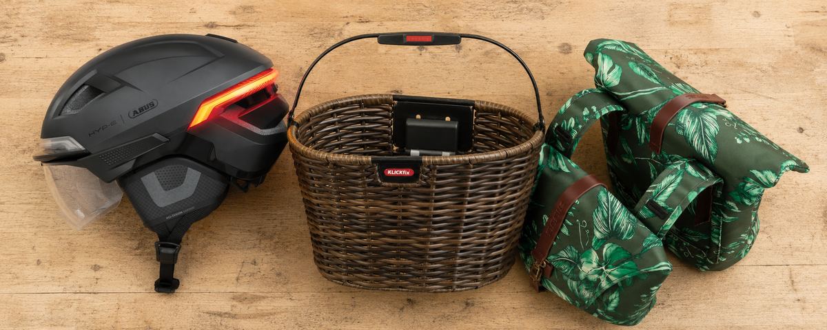 Trois accessoires posés sur un établi en bois : un casque ABUS à feu arrière intégré, un panier avant en osier et une paire de sacoches à motif feuillage