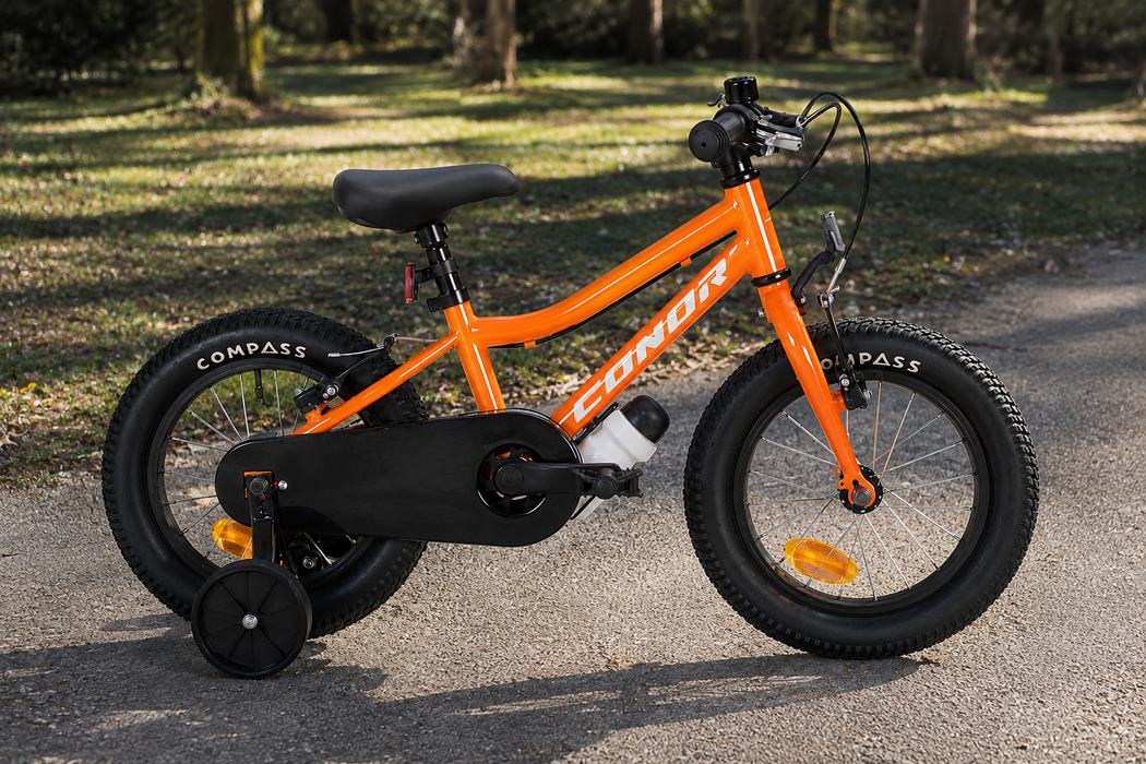 Vélo enfant Conor 14 pouces orange, sur ses roulettes d'apprentissage, dans une allée de parc